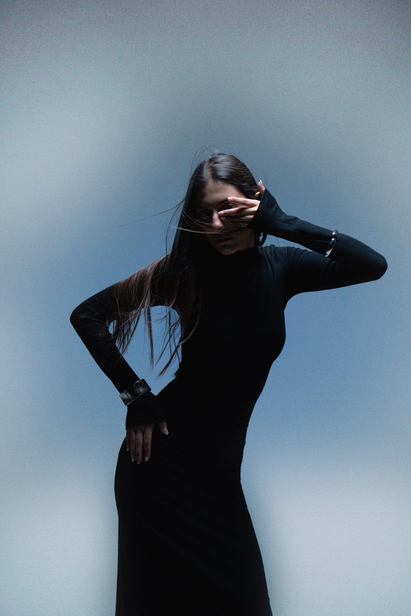 Body, Blur, Flash: mavi fonda siyah elbiseli manken, bir eli gözünde, dirseği havada